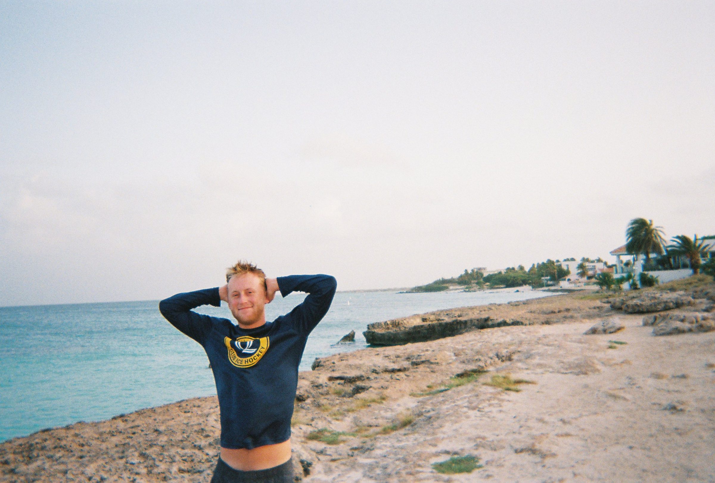 Ty smiling with his hands behind his head on a rocky shoreline beside turquoise water